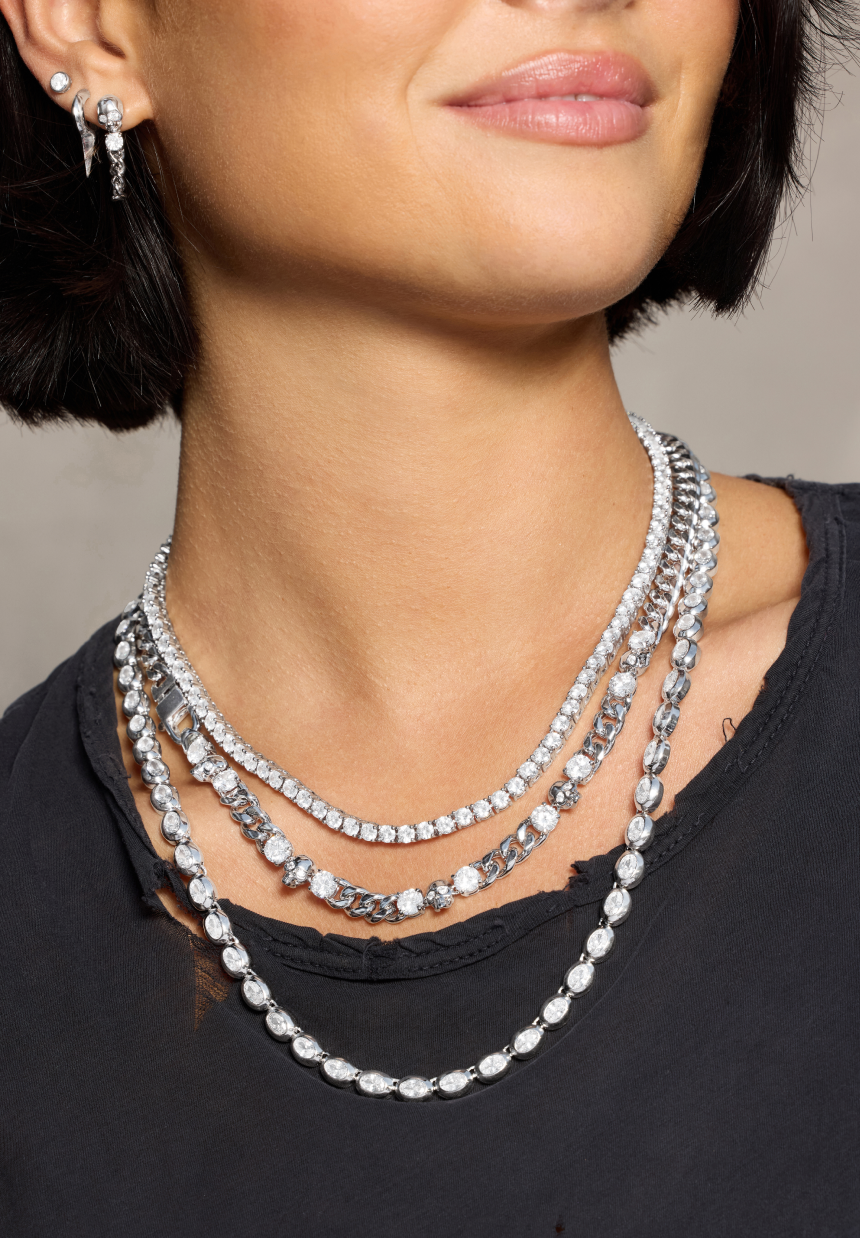 Close-up of a woman wearing multiple silver necklaces and earrings against a neutral background