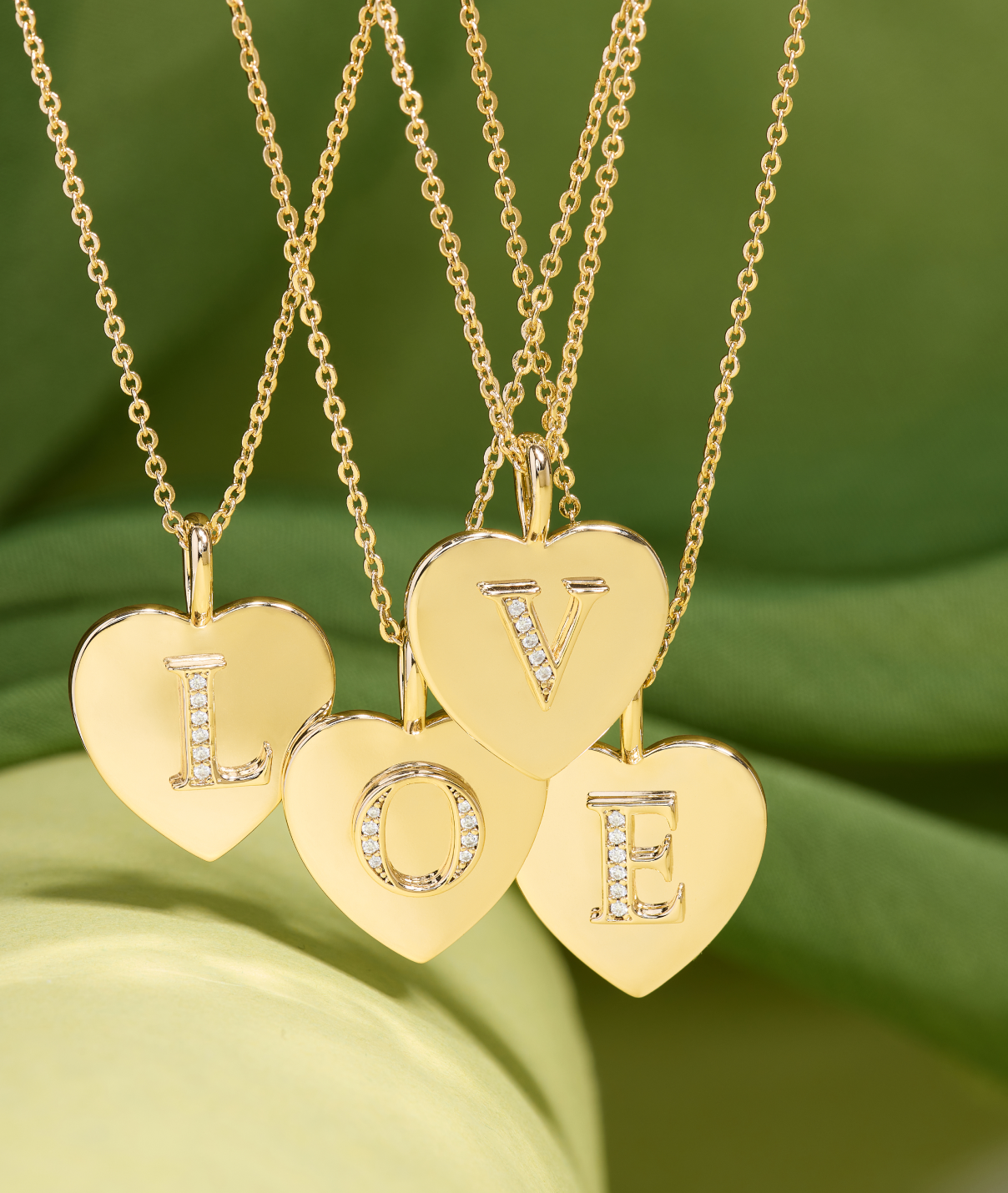 Gold necklaces with heart-shaped pendants spelling 'LOVE' on a green leaf background