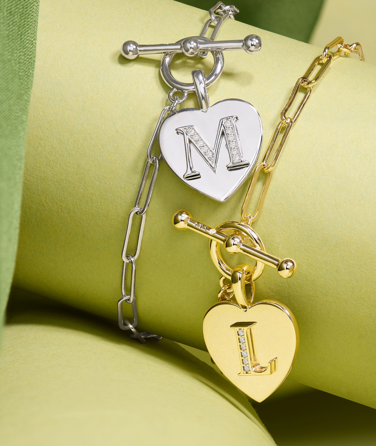 Two bracelets, one silver and one gold, with heart-shaped charms featuring initials on a green background.