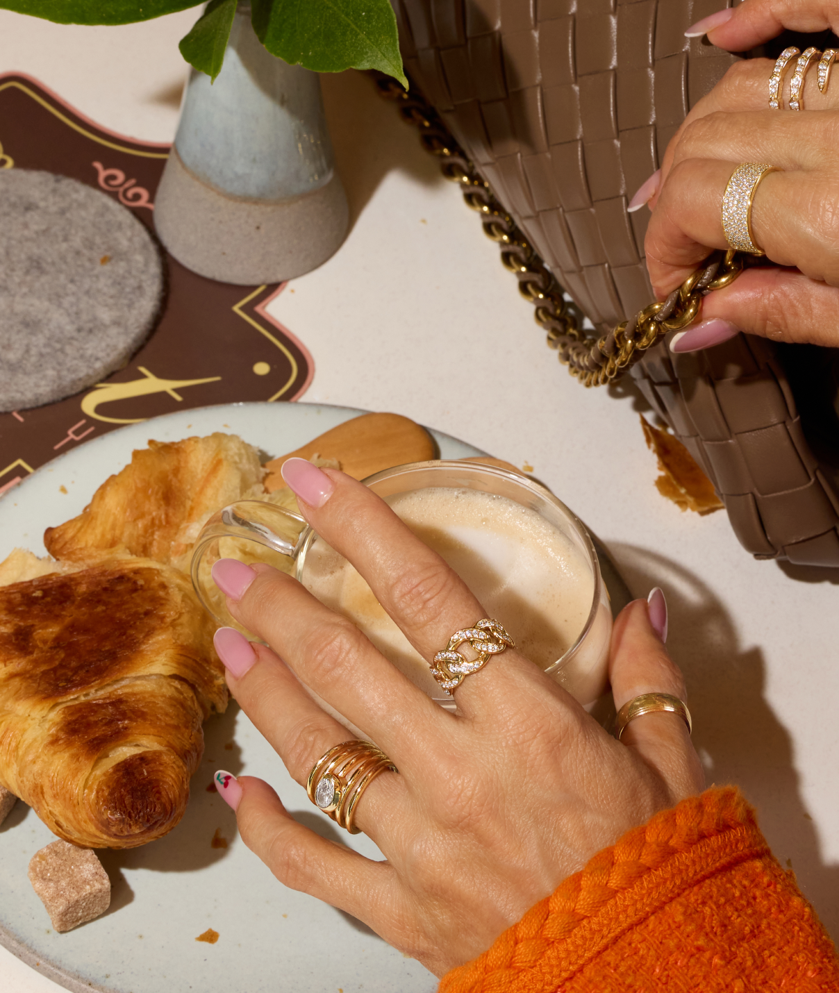 Person holding a glass of coffee with croissants on a plate, wearing multiple rings.