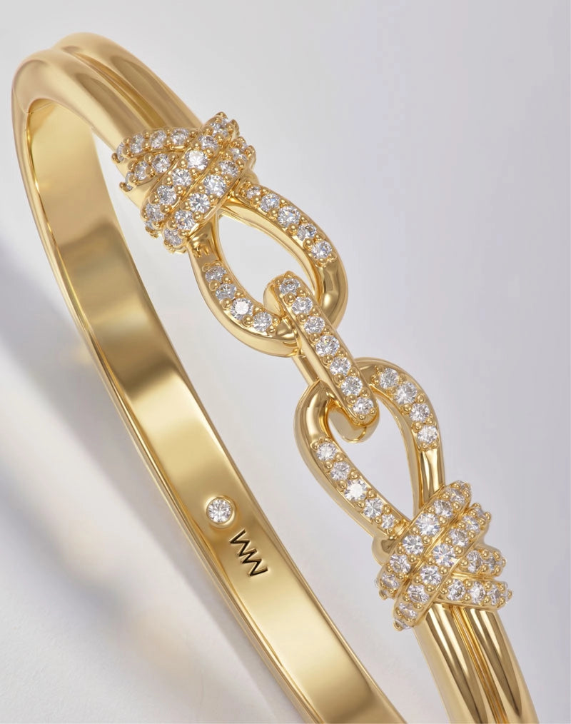 A close-up of a gold bangle bracelet featuring three interlocking knot motifs paved with small clear stones, shown on a light gray background. The inner band is engraved with the brand name 'MM'.