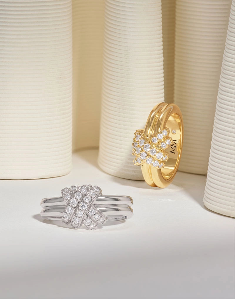 Two knotted ring designs displayed on a white surface with cream ribbed cylindrical props in the background. One ring is silver-toned with pavé-set clear stones, and the other is yellow gold with a similar stone cluster and an 'MM' mark inside the band.
