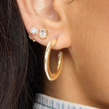 A close-up of a person's ear wearing a polished gold huggie hoop earring with a small clear gemstone accent, alongside matching stud earrings.