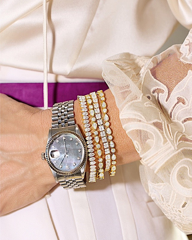 Silver watch on a wrist with multiple gold bracelets, wearing a white lace sleeve.