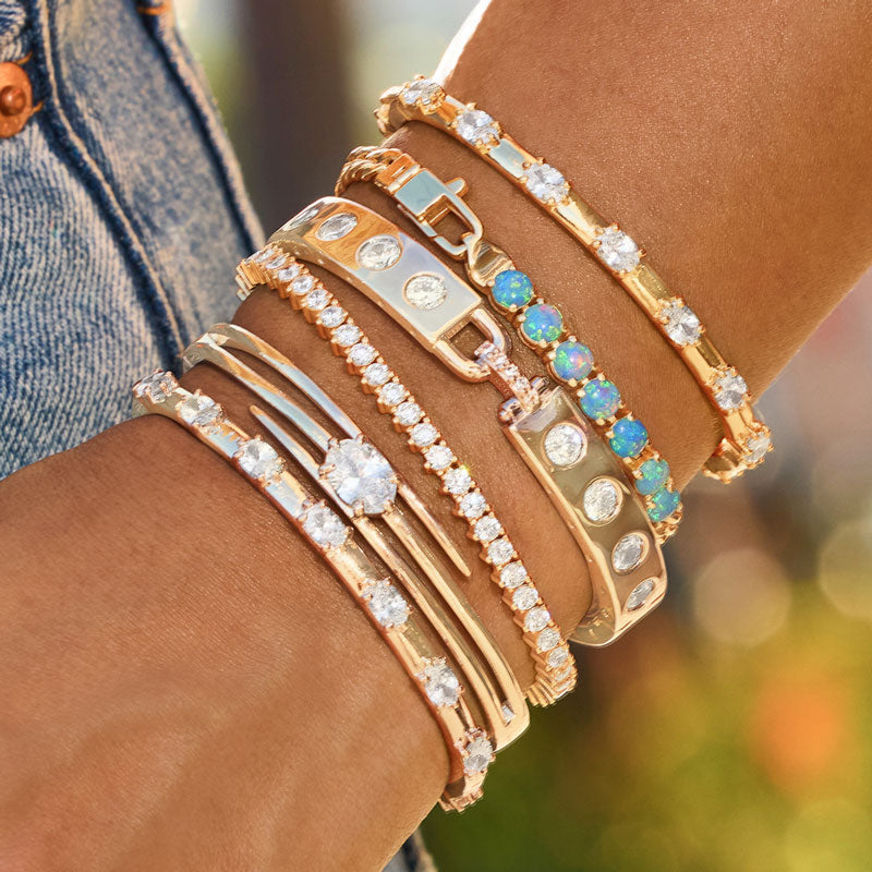 Close-up of a wrist wearing multiple gold and rose gold bracelets with gemstones against a blurred background.
