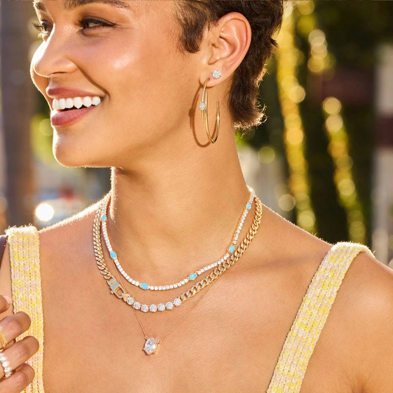 A close-up lifestyle shot of a woman wearing layered gold jewelry, including a double-strand necklace with white pearls, turquoise beads, and a gold box charm, a gold hoop earring with a pearl stud, and a delicate gold pendant necklace.