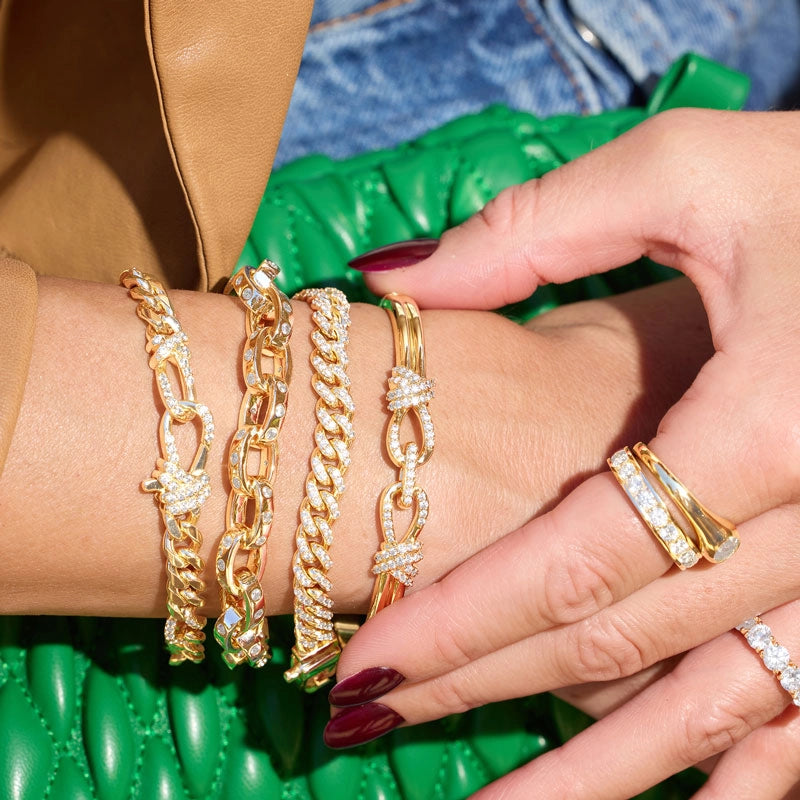 A close-up of a person's wrist and hand wearing multiple gold chain bracelets, including curb links, rope links, and knot-style bracelets with clear stones, alongside gold rings, against a green quilted bag and denim background.