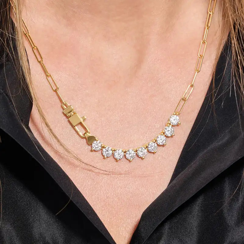 A close-up of a gold chain necklace with a row of clear gemstones, worn on a model's neck over a black satin top.