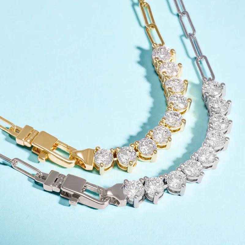 Two tennis-style necklaces with round-cut clear stones, one in yellow gold and one in silver, arranged on a light blue background.