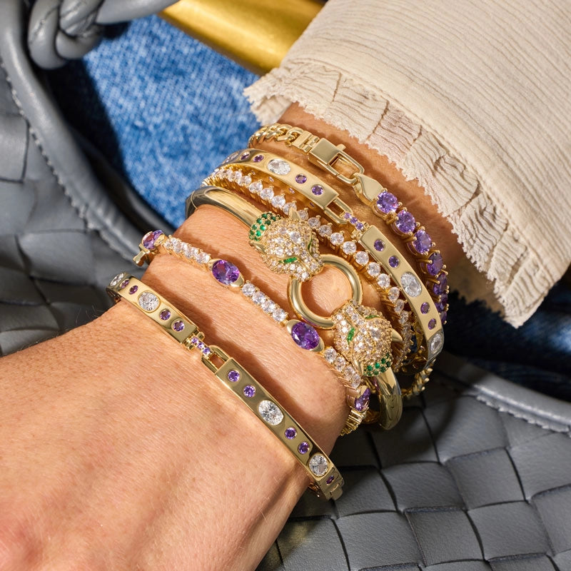 A close-up of a wrist wearing a stack of gold and silver bracelets adorned with purple, white, and green gemstones, resting against a gray woven leather bag.