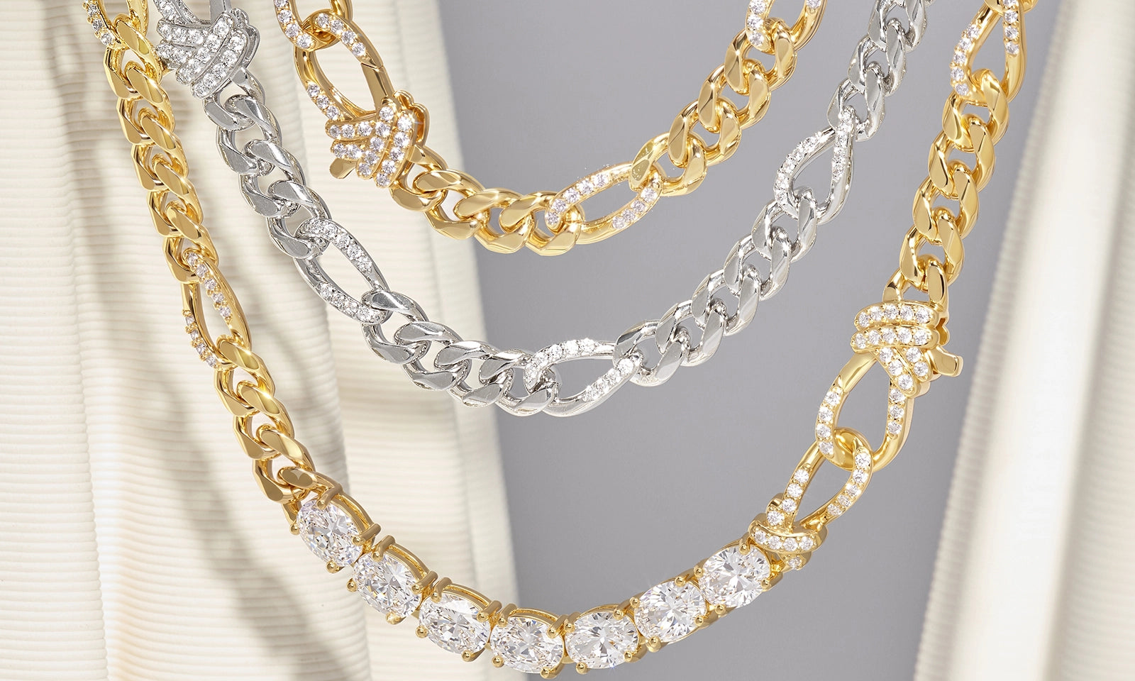 Four layered chain necklaces displayed on a cream-colored jewelry stand. The necklaces include gold and silver-tone links, with some pieces featuring pavé-set clear stones.