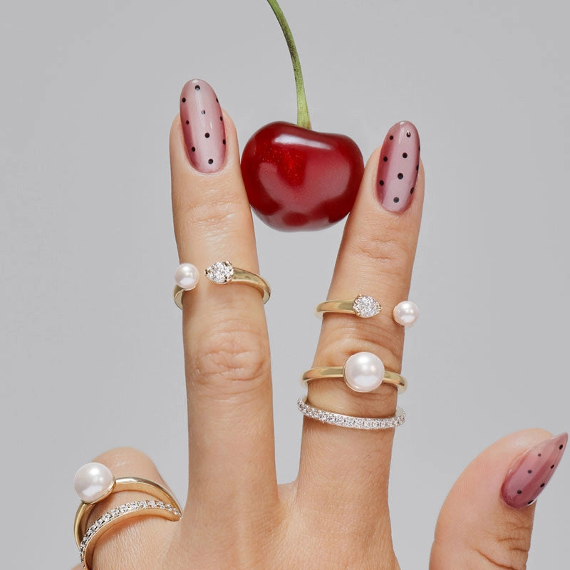 A close-up of a hand with pink dotted nail art wearing multiple gold and silver rings with pearl and crystal details, holding a red cherry against a light gray background.