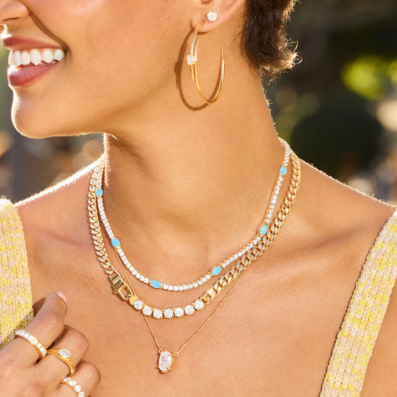 Woman wearing gold hoop earrings and multiple necklaces with a blurred outdoor background