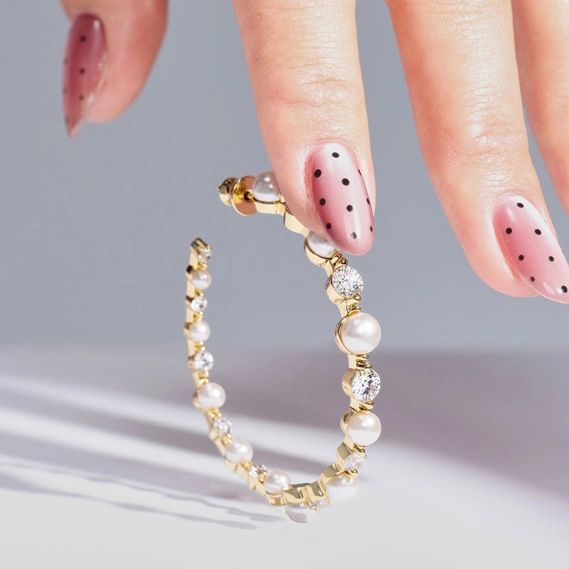 Gold hoop earrings adorned with white pearls and clear rhinestones, held by a hand with pink nail art featuring black polka dots, against a light gray background.