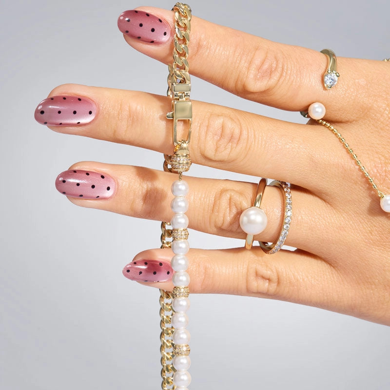 A close-up of a hand displaying a gold chain necklace with white pearl beads and matching gold rings with pearl details against a light gray background.
