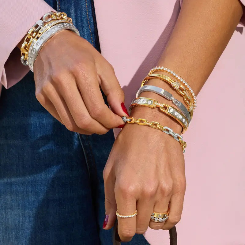 A close-up of a person's hands and wrists wearing multiple gold, silver, and white gold bracelets and rings against a soft pink background.