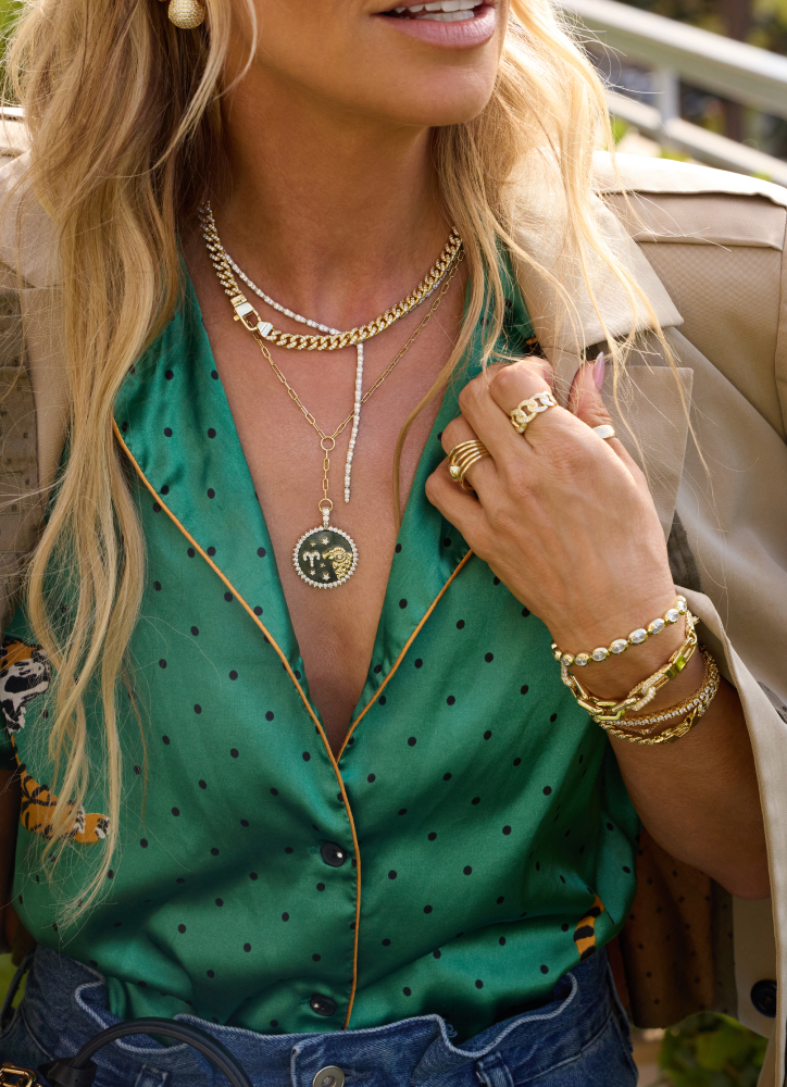 Woman wearing gold jewelry including necklaces, bracelets, and rings, with a green top and beige jacket.