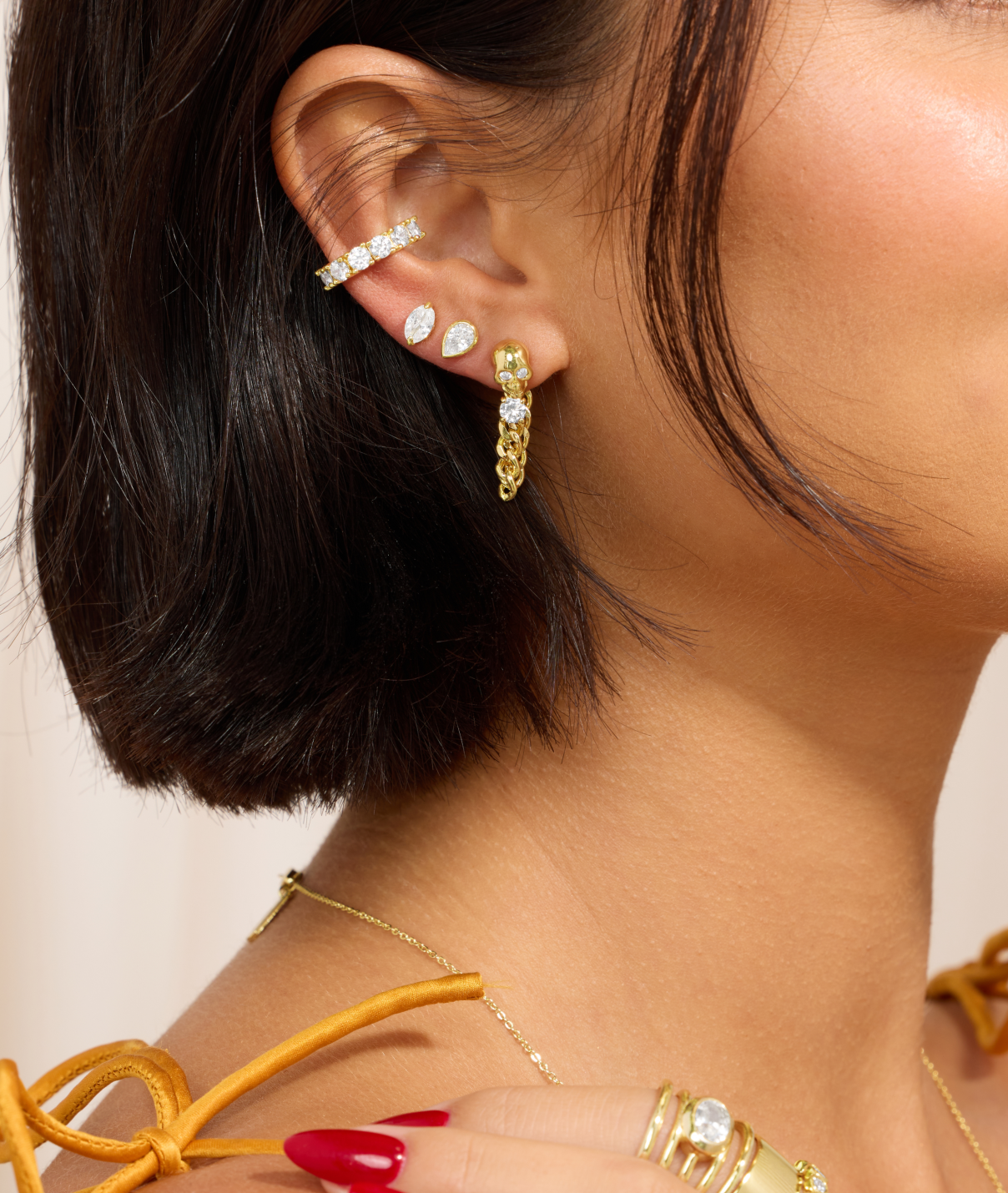 Close-up of a person wearing gold hoop earrings and multiple gold rings on a neutral background